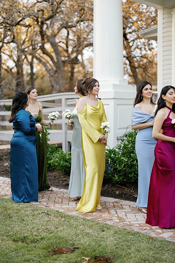 Bridesmaid portraits of a lineup holding mini white bouquets in colorful dresses on a porch with white columns and fall trees behind