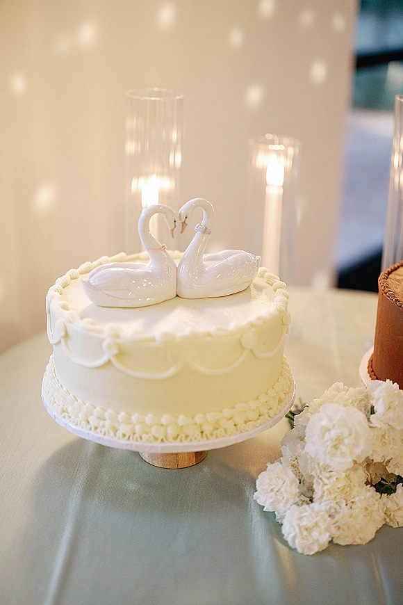 Wedding cake with scalloped buttercream, a swan topper, and candles on a pedestal stand beside white flowers against a light wall