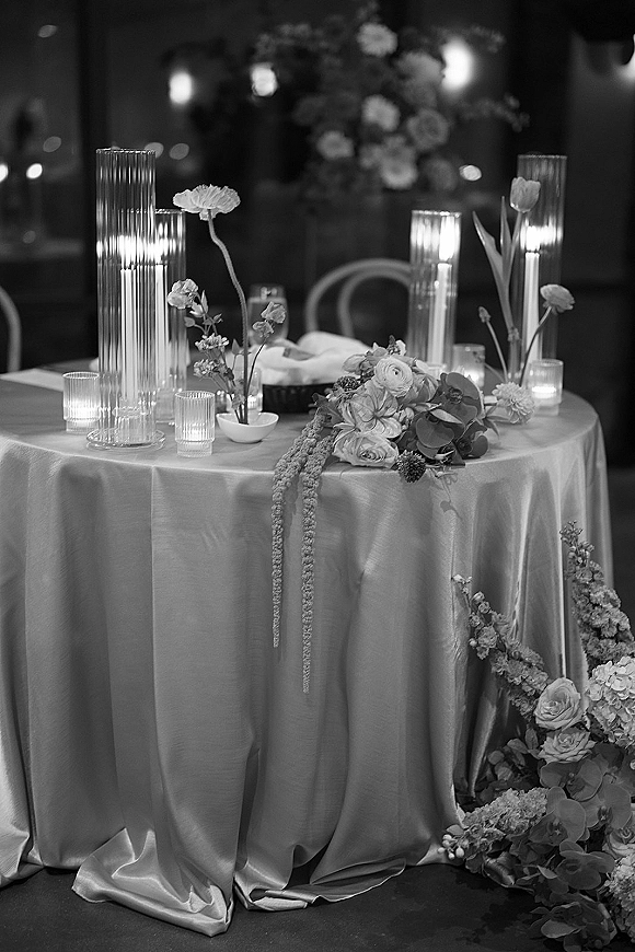 Reception tablescape with satin tablecloth, floral centerpiece and taper candles in ribbed glass holders, glowing in a dim reception room