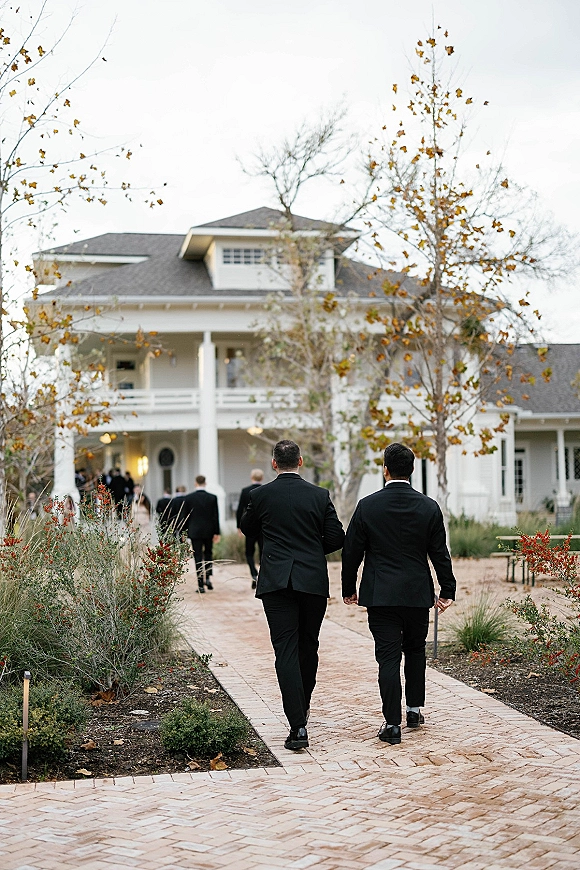 Wedding guests arriving in black suits walk up a brick walkway toward a large white house, framed by autumn shrubs and pathway lights