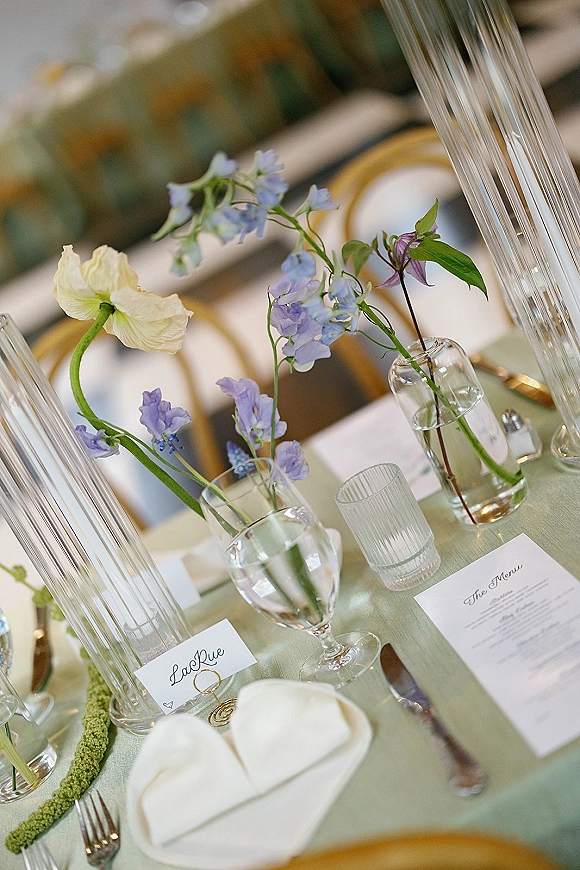 Reception tablescape with wedding table decor, blue and white blooms in bud vases and tall glass cylinders on a green tablecloth with gold-rim plates.