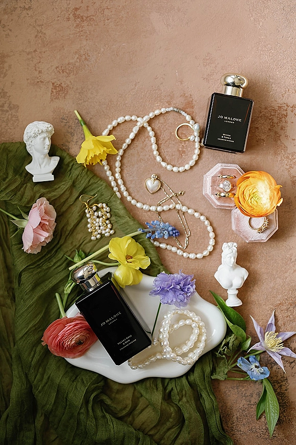 Wedding details flatlay with wedding rings flatlay, pearl necklace and earrings, perfume bottles, blooms and green drape on stone surface