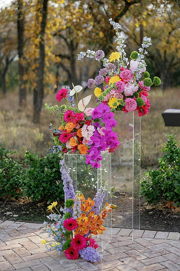 Wedding floral installation with acrylic pedestal wedding flowers in bright orchids and roses along a stone paver garden walkway under trees