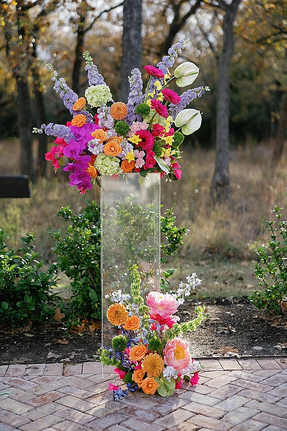 Wedding floral arrangement on a clear acrylic pedestal with roses, orchids, and hydrangea in a garden along a brick walkway