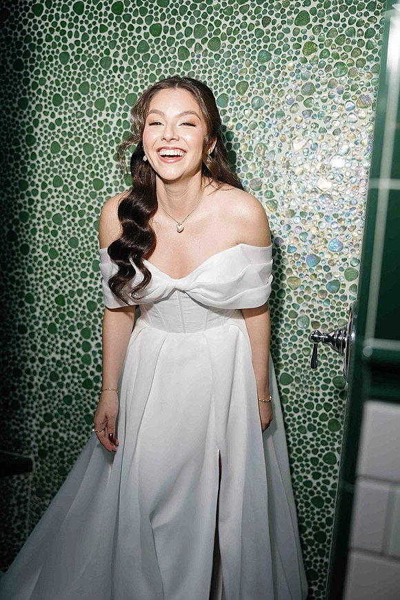Bridal portrait of a laughing bride in an off the shoulder wedding dress with pearl earrings, posed by a green mosaic tile wall