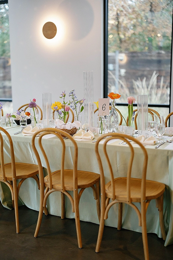 Reception tablescape with wedding head table decor featuring wildflower centerpieces and bud vases on a pastel tablecloth by large windows