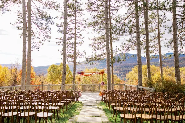 Outdoor ceremony setup with wood ceremony chairs lining a stone aisle to a draped wooden arch, framed by pine trees and mountain views