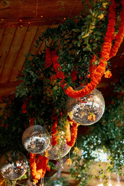 Hanging reception decor with disco ball wedding decor, marigold garlands and greenery beneath a wood ceiling with string lights among trees
