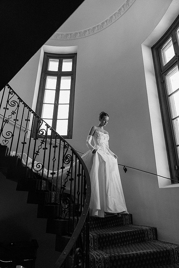 Bridal portrait of a bride on staircase in an off-the-shoulder gown and long gloves, lit by tall windows beside a wrought iron railing