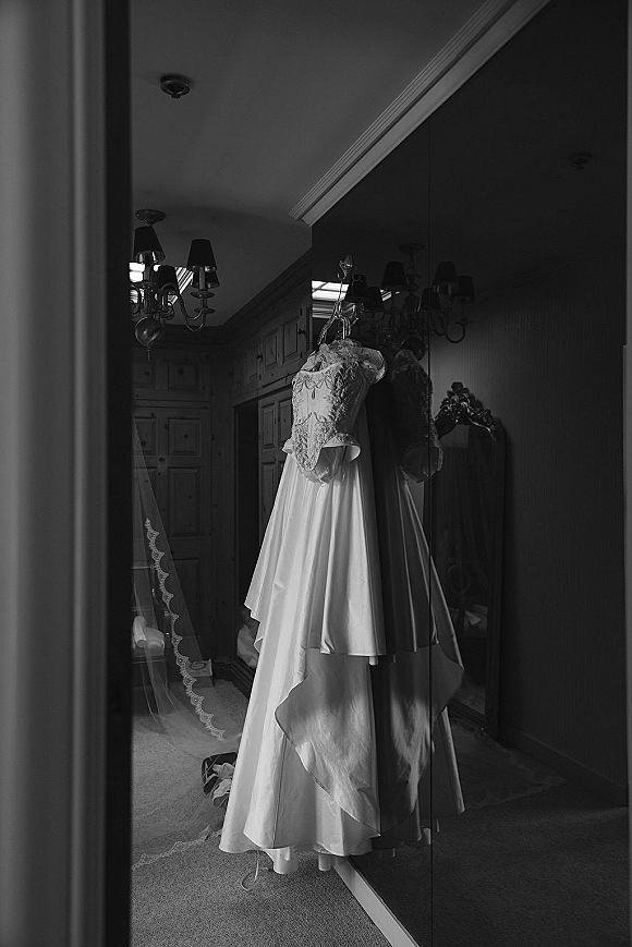 Wedding dress with a lace bodice hanging on a hanger, satin skirt draping in a hallway with mirrored closet doors reflecting it.