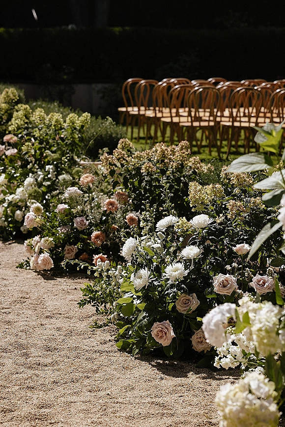 Ceremony aisle florals with blush roses, hydrangeas and greenery along a gravel path beside wooden chairs in a garden setting