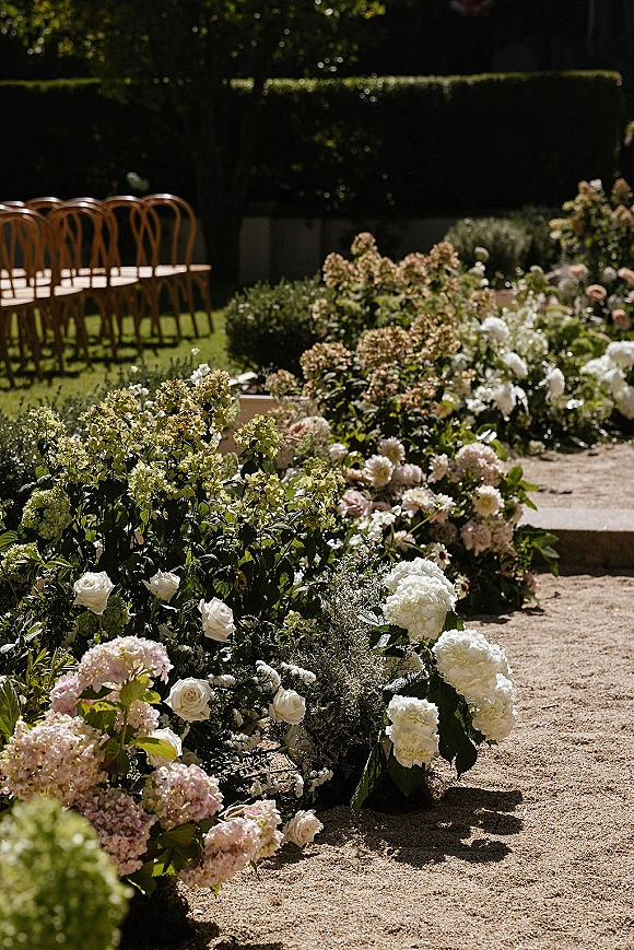 Ceremony aisle flowers with ground floral aisle arrangements of white roses, hydrangea, and greenery along a gravel garden path with wooden chairs