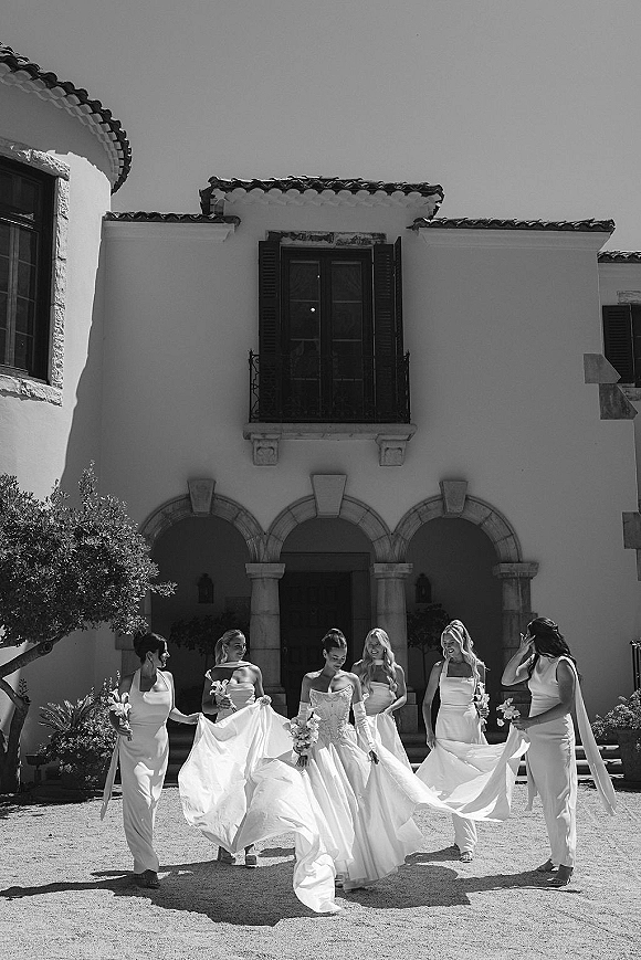 Bride with bridesmaids walking in a villa courtyard, bouquets with ribbon streamers as friends lift her full skirt gown