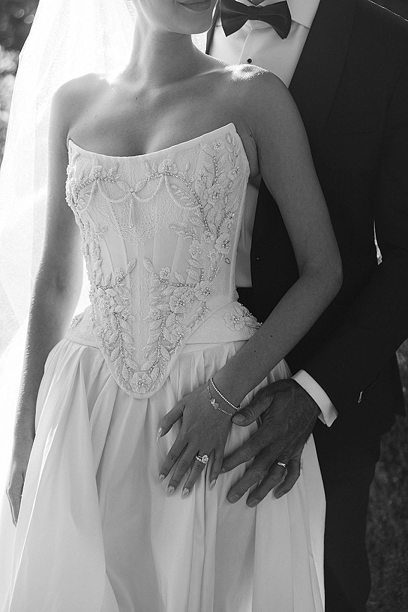 Wedding couple portrait of groom hugging bride, highlighting their hands with wedding rings, lace bodice and veil in sunlit greenery backdrop