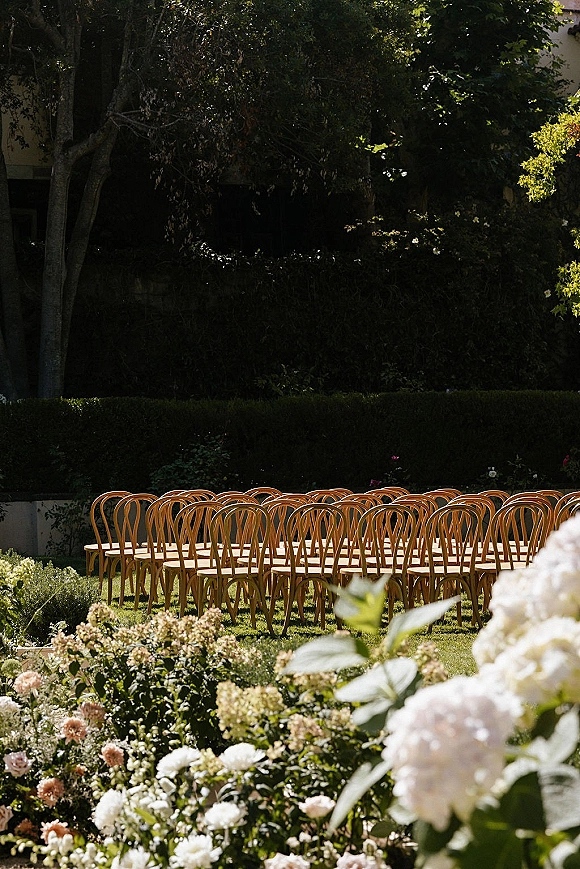 Ceremony seating with outdoor wedding chairs in neat rows, wood accents and garden flowers along hedges on a sunlit lawn by trees