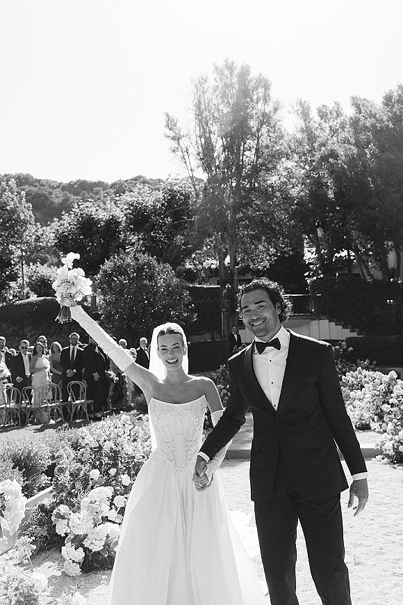 Recessional moment as bride and groom walk hand in hand, bride raising bouquet, veil flowing down a sunlit garden aisle with guests behind