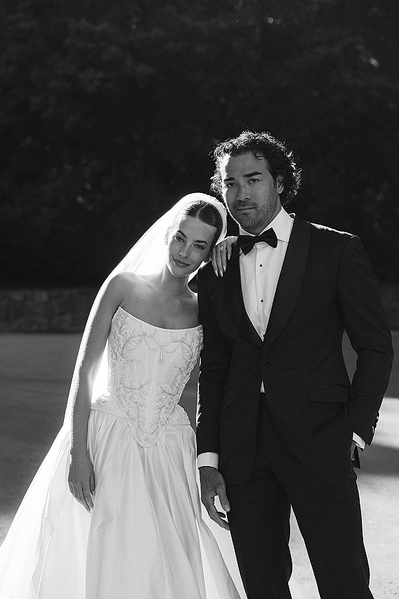 Couple portrait of bride leaning on groom, showing her bridal veil and beaded strapless dress in sunlit trees and lawn backdrop