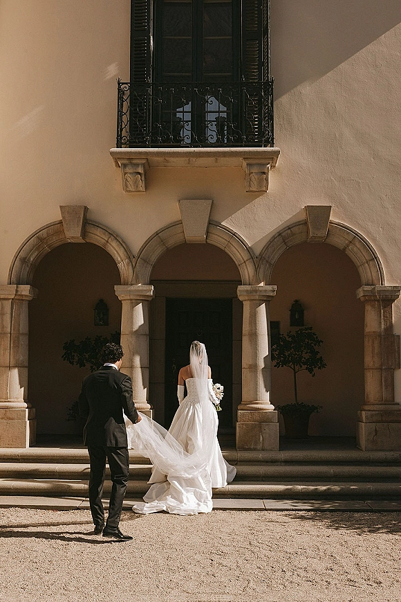 Couple portrait from behind as bride and groom walk away, groom holding veil, with cathedral train and calla lily bouquet in stone colonnade courtyard.