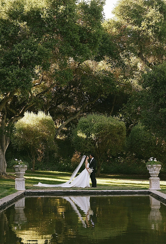 Couple portrait of bride and groom by pool, facing each other as her veil train flows, holding a bouquet on an estate garden lawn