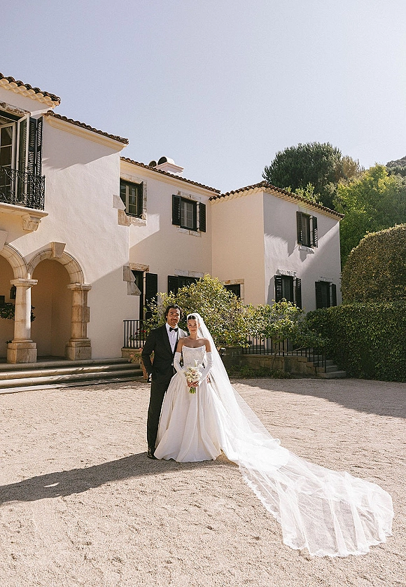 Couple portrait of bride in a strapless gown with long veil and bouquet beside groom in tuxedo before a stucco villa courtyard doorway