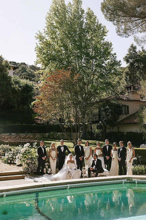 Wedding party portrait of bride and groom with friends beside a pool, bride in veil holding bouquet amid black-tie bridesmaids and groomsmen