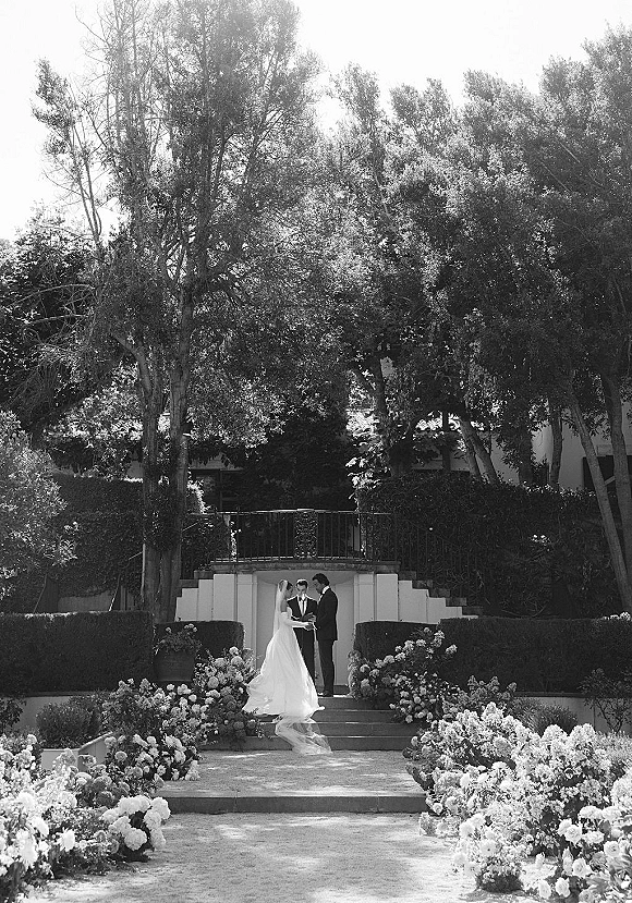 Wedding vows as bride in veil and groom in black suit face the officiant on garden steps, with floral arrangements and hedges behind