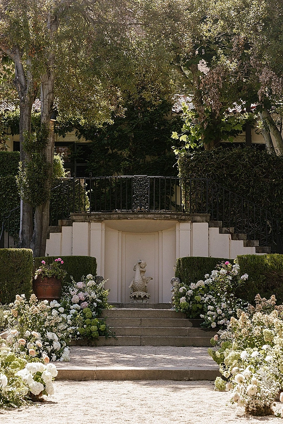 Ceremony aisle decor with a floral lined aisle of hydrangeas and roses on stone steps, accented by a garden statue and villa balcony backdrop