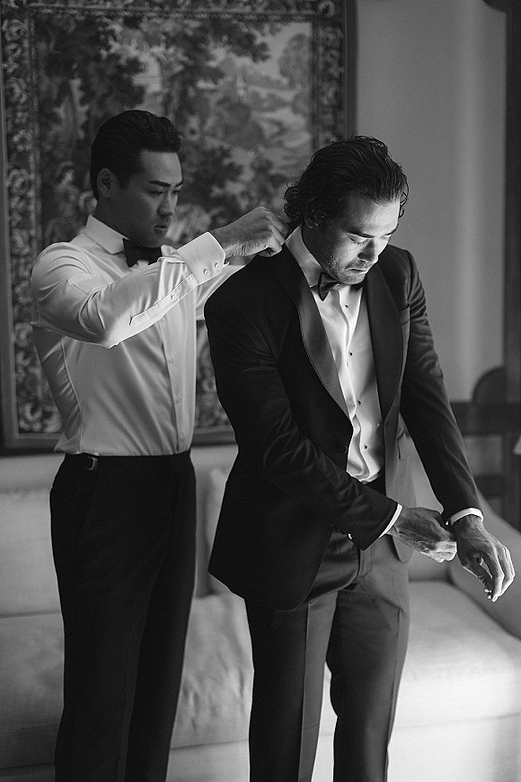 Groom getting ready as groomsmen help him put on his tuxedo jacket, adjusting a bow tie in a softly lit hotel room