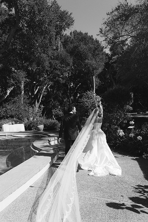 Wedding couple portrait of bride and groom holding hands walking away, her long veil trailing as they stroll by a garden fountain path