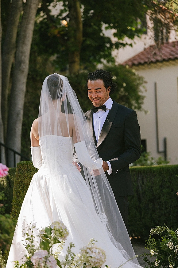 First look moment as bride in a long veil and white opera gloves meets groom in black tuxedo amid sunlit garden greenery and stucco building