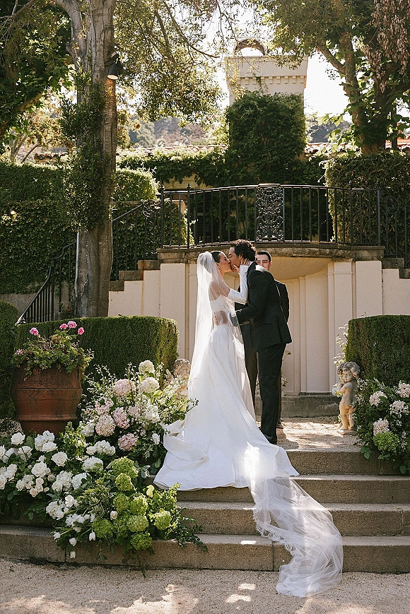 Wedding kiss as bride and groom embrace on stone steps, long veil trailing among white hydrangea and rose florals on a terrace