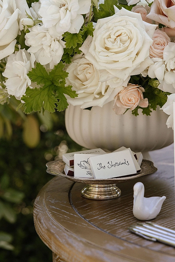 Wedding escort cards with calligraphy escort cards on a silver compote, framed by garden roses and hydrangea on a rustic wood table outdoors