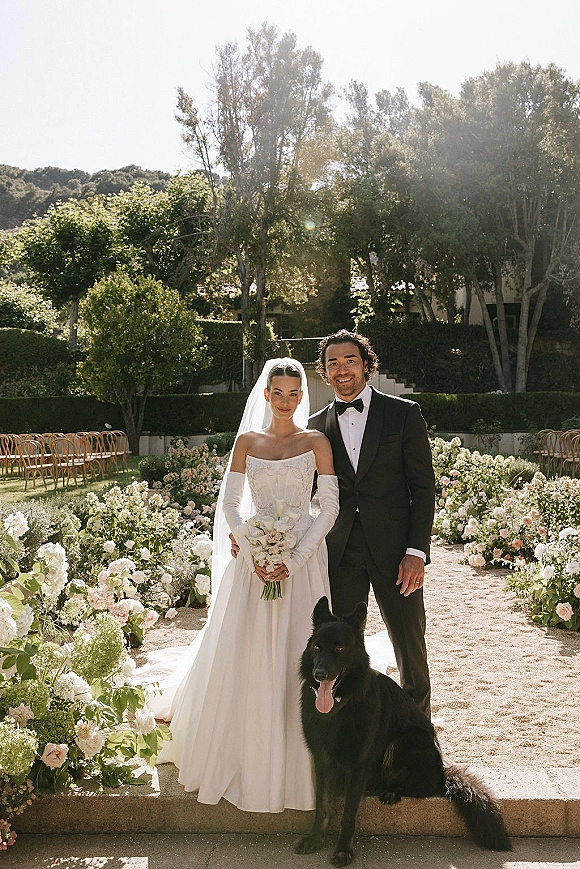 Couple portrait of bride and groom with dog, bride in strapless gown and veil holding bouquet beside sunlit garden aisle chairs