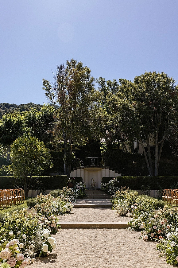 Ceremony aisle design with a floral lined aisle of white and blush ground blooms and greenery leading to stone steps in a garden setting