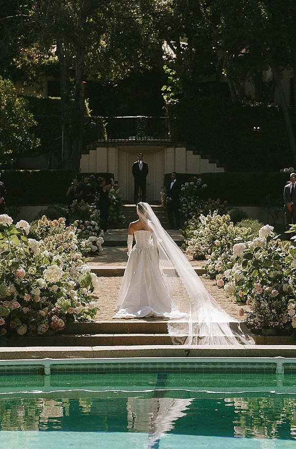 Ceremony processional as bride walking down aisle in a strapless gown with long veil and opera gloves, garden steps lined with flowers