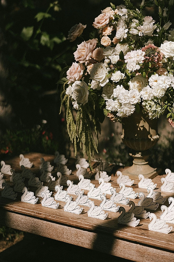 Wedding escort cards with swan escort cards set on a wooden table beside an urn of blush roses and white blooms in dappled garden light