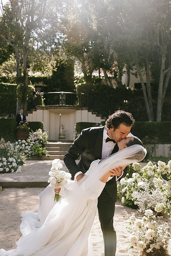 Wedding kiss portrait of groom dipping bride as they kiss under her veil, bouquet of white florals and greenery on sunlit garden steps