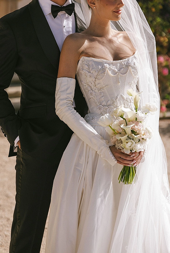 Couple portrait of bride and groom portrait on an outdoor walkway, groom behind bride as she holds a calla lily bouquet amid greenery