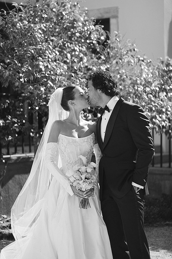 Wedding kiss as bride and groom embrace, her veil flowing and bouquet in hand, in a garden with trees and a fence backdrop
