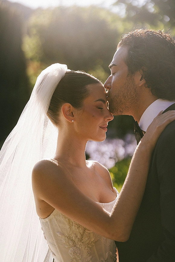 Wedding couple portrait with a forehead kiss, bride in a veil and strapless lace dress beside groom in tuxedo in sunlit greenery