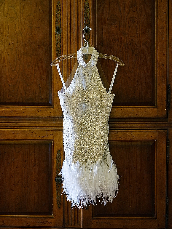 Wedding dress with sequin wedding dress sparkle, halter neckline and feather hem hanging on a clear hanger against a carved wooden wardrobe