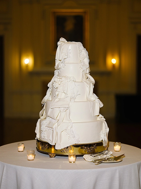 Wedding cake with white fondant and lace appliqué on a gold cake stand, surrounded by votive candles in a dim reception room