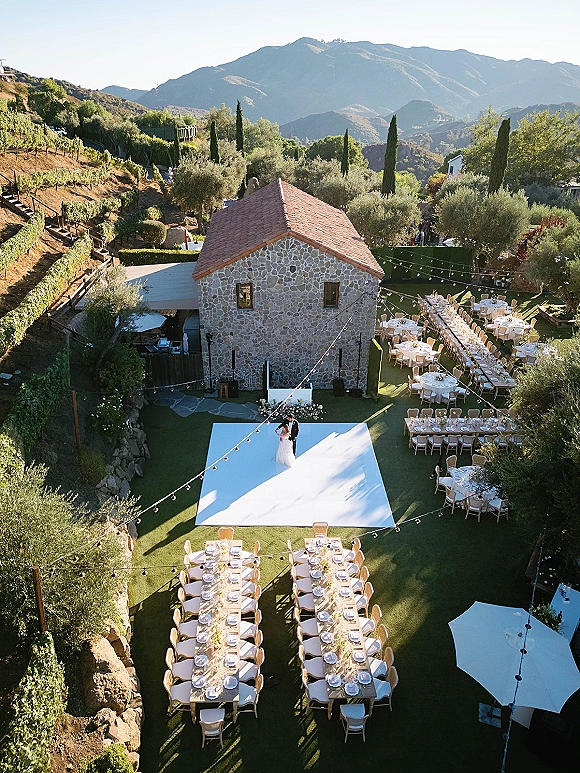 Outdoor reception setup with al fresco wedding reception tables under string lights, white dance floor on lawn by stone cottage and vineyard hills.