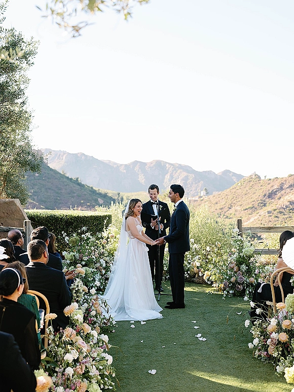 Wedding vows during an outdoor wedding ceremony as bride and groom hold hands before officiant on a flower-lined aisle with mountains behind