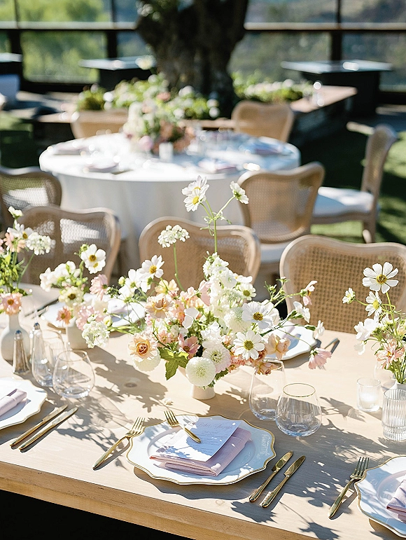 Reception tablescape with wildflower centerpieces, blush napkins, gold-rim plates and flatware, and votive candles on an outdoor lawn under a large tree