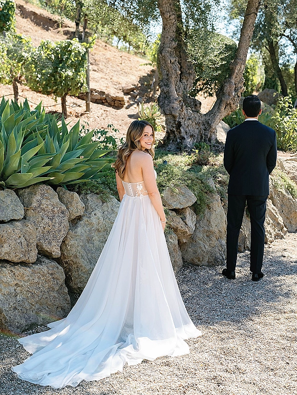 Wedding first look as the bride approaches the groom from behind, her lace bodice and tulle train flowing on a gravel path by a stone wall