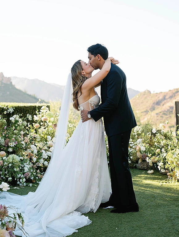 Wedding kiss portrait of bride and groom kissing, her cathedral veil and long train flowing on a garden lawn with mountain view backdrop