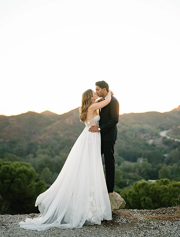 Wedding kiss portrait of bride and groom kissing, her lace gown train flowing as they embrace on a rocky mountain overlook