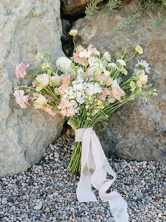 Bridal bouquet of pastel wedding bouquet flowers tied with a white ribbon, resting on rocks and gravel with greenery in the background