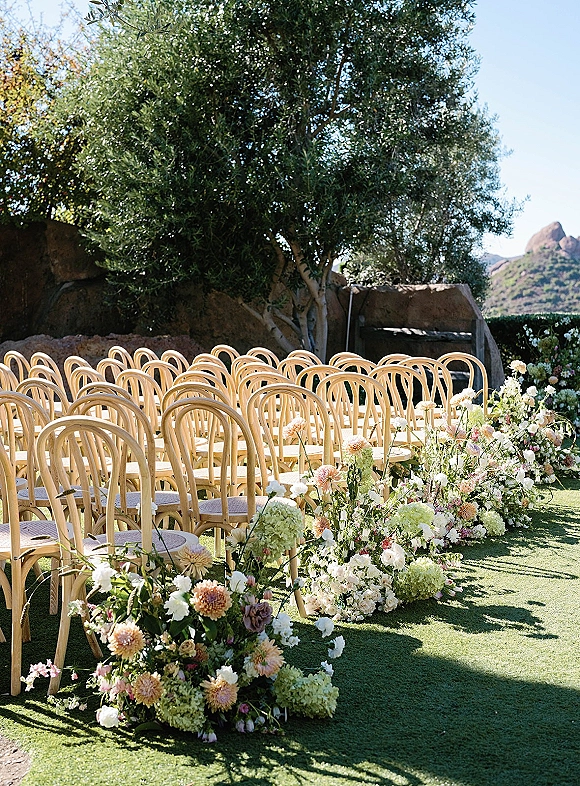 Ceremony aisle decor with a ground floral aisle of hydrangeas, dahlias and roses beside wood chairs on a lawn with mountain backdrop